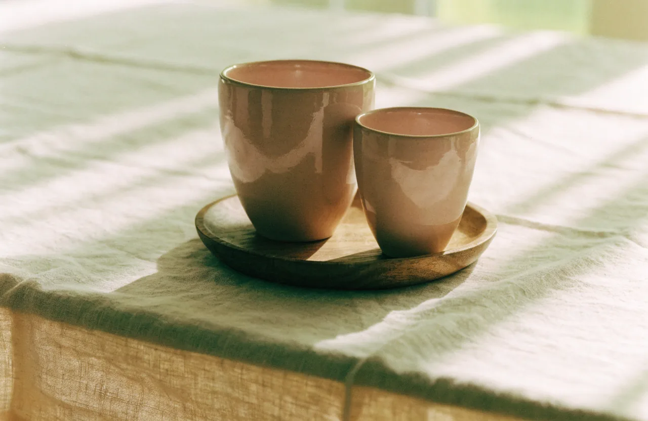 Two dusty rose ceramic cups, one larger and one smaller, standing together on a small wooden tray on warm linen.
